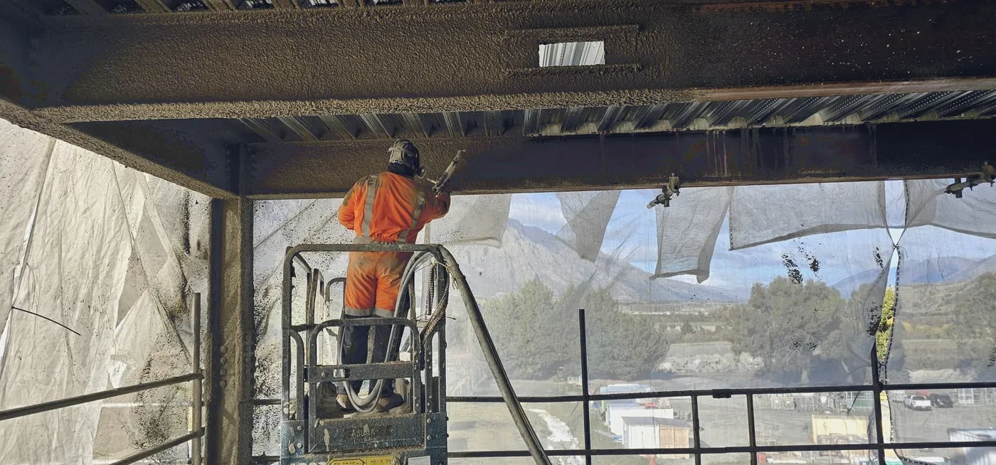 Applicator spraying cementitious protection to a beam from an elevated work platform